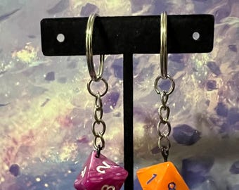 Puede incluir: Dos llaveros con forma de dados, uno es morado y naranja con números blancos, el otro es naranja con números blancos. Ambos dados cuelgan de cadenas plateadas.