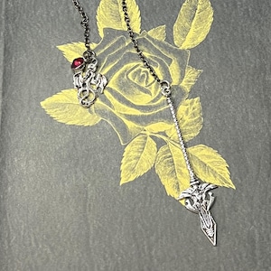 May include: A silver-toned pendant with a chain, featuring a dragon charm with a red gem and a decorative sword-like charm. The pendant hangs against a gray background with a yellow rose illustration.