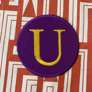 May include: A purple felt circle patch with a gold embroidered letter U.