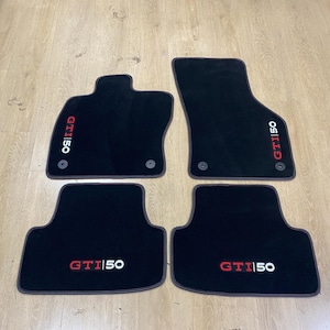 Puede incluir: Juego de cuatro alfombrillas negras para coche. Cada alfombrilla tiene el logotipo "GTI 50" en rojo y blanco. Las alfombrillas tienen un borde gris oscuro y están diseñadas para el interior de un vehículo. Fabricadas con material suave y afelpado.