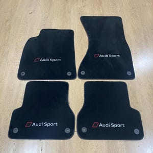 以下が含まれることがあります： 白と赤で「Audi Sport」の文字が入った黒い車のフロアマット4枚。マットは明るい木製の表面に配置されています。各マットには円形の留め具が付いています。