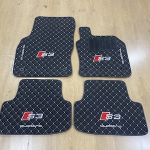 Puede incluir: Un juego de cuatro alfombrillas negras para coche con un patrón de costuras de diamantes en blanco. Cada alfombrilla presenta un logotipo S3 en rojo y blanco y la palabra quattro en texto blanco. La alfombrilla del conductor incluye un refuerzo.