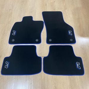 May include: Set of four black car floor mats with blue trim. Each mat features a white and blue "R" logo. The mats are designed to fit a vehicle's interior and protect the floor from dirt and wear.