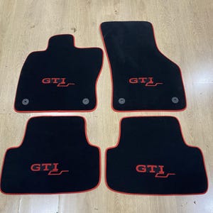 May include: A set of four black automotive floor mats with red piping edges. Each mat features the red embroidered text GTI. The two front mats include two circular mounting grommets each, while the two rear mats have a rectangular shape.