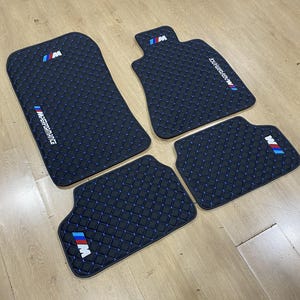 Può includere: Un set di quattro tappetini neri per auto con motivo a rombi trapuntato e filo blu. Ogni tappetino mostra il logo M in blu, rosso e bianco, con due tappetini che presentano anche la scritta bianca M PERFORMANCE stampata sulla superficie.