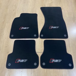 May include: Set of four black car floor mats. Each mat has a red and white "RS7" logo. The mats are shaped to fit a vehicle's floorboards and have circular fasteners. The mats are made of a soft, dark material.
