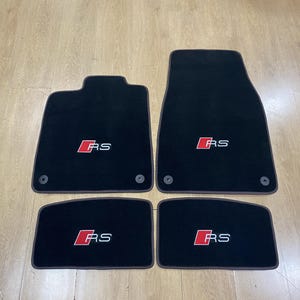 May include: A set of four black car floor mats with dark brown edging. Each mat displays a red square emblem containing the white text RS. The two larger front mats feature circular fixing points, and the two smaller rear mats are rectangular in shape.