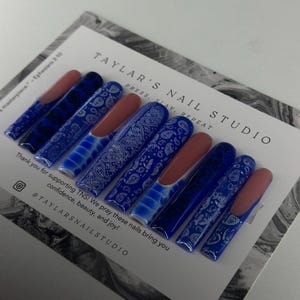 May include: A set of blue and white press-on nails with a paisley pattern. The nails are long and have a square shape. The text "TAYLAR'S NAIL STUDIO" is printed on the card. The text "PRESS. SLAY. REPEAT" is printed on the card. The text "Thank you for supporting TNS! We pray these nails bring you confidence, beauty, and joy!" is printed on the card. The text "@TAYLARSNAILSTUDIO" is printed on the card.