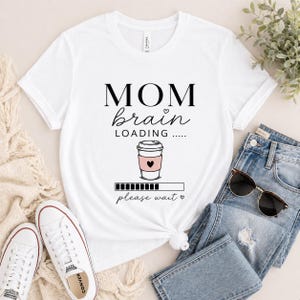May include: White t-shirt featuring the text "MOM brain LOADING....." above a coffee cup graphic with a pink heart and a loading bar. The shirt is styled with blue jeans and white trainers.