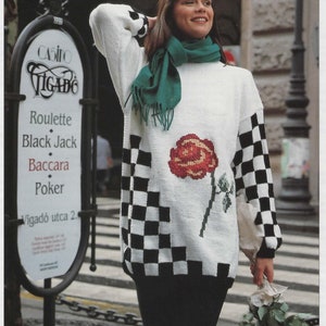 May include: A white sweater with a black and white checkered pattern on the sleeves and a red rose embroidered on the front. The sweater is worn by a woman with long brown hair and a green scarf.