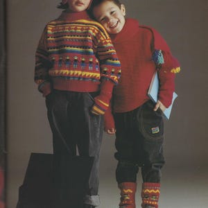 May include: Two children wearing knitted winter clothing. One child wears a red hat, sweater, and matching mittens with a colorful pattern. The other child wears a red turtleneck sweater, dark jeans, and red socks with a yellow and blue pattern.
