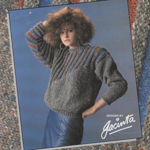 May include: A gray sweater with blue, red, and brown stripes on the shoulders and sleeves. The sweater has a round neckline and is knitted in a textured stitch. The model is wearing a blue leather skirt and has curly brown hair. The image includes the text "Designs by Jacinta".