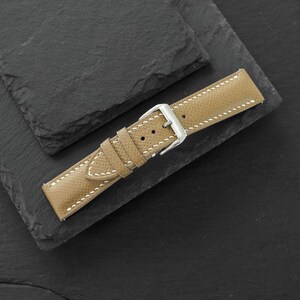May include: A tan leather watch strap featuring a textured surface, white contrast stitching, and a polished silver-tone metal buckle. The strap includes two matching leather keepers and multiple adjustment holes for securing the watch band.