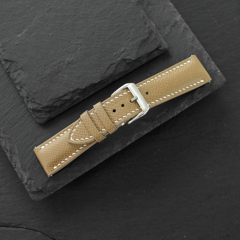 May include: A tan leather watch strap featuring a textured surface, white contrast stitching, and a polished silver-tone metal buckle. The strap includes two matching leather keepers and multiple adjustment holes for securing the watch band.