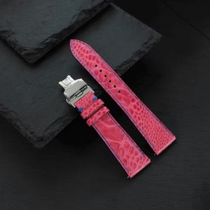 Pode incluir: Uma pulseira de relógio rosa brilhante com superfície texturizada e costura azul. A pulseira possui um fecho dobrável de metal prateado e dois passadores de couro rosa a condizer. O design inclui uma série de furos pré-perfurados.