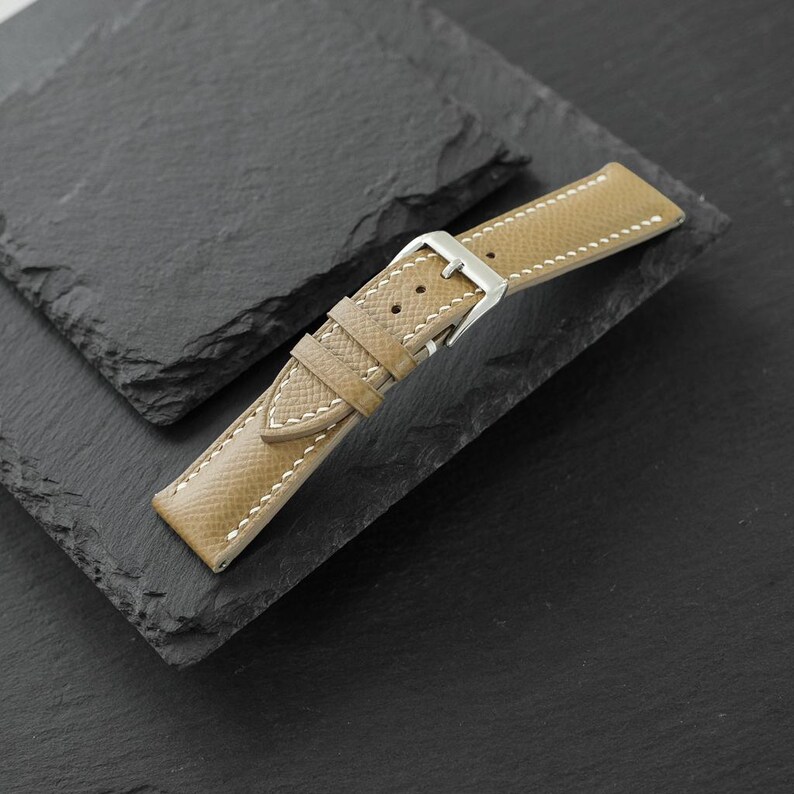May include: A tan leather watch strap featuring a textured surface, white contrast stitching, and a polished silver-tone metal buckle. The strap includes two leather keepers and multiple adjustment holes for securing the watch to a wrist.