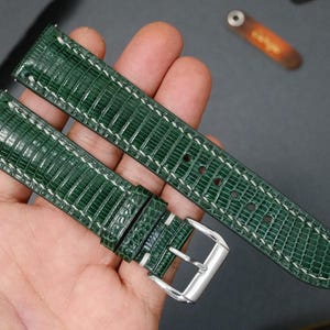 Puede incluir: Correa de reloj verde con un patrón de piel de lagarto texturizado y costuras blancas. La correa tiene una hebilla plateada y se muestra sobre un fondo oscuro.