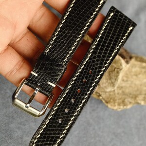 Puede incluir: Una correa de reloj negra con un patrón texturizado de piel de lagarto y costuras blancas en contraste. La correa cuenta con una hebilla de metal plateado pulido y múltiples orificios de ajuste, mostrando un diseño clásico y refinado.