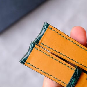 Op de afbeelding: Een horlogeband met een donkergroene buitenkant met krokodillenstructuur en een gladde mosterdgele binnenvoering. De band is afgewerkt met zichtbare donkergroene stiksels langs de randen en bevat voorgeponste gaten voor het verstellen.