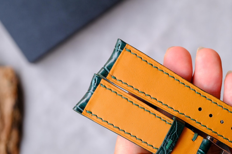 Op de afbeelding: Een horlogeband met een donkergroene buitenkant met krokodillenstructuur en een gladde mosterdgele binnenvoering. De band is afgewerkt met zichtbare donkergroene stiksels langs de randen en bevat voorgeponste gaten voor het verstellen.
