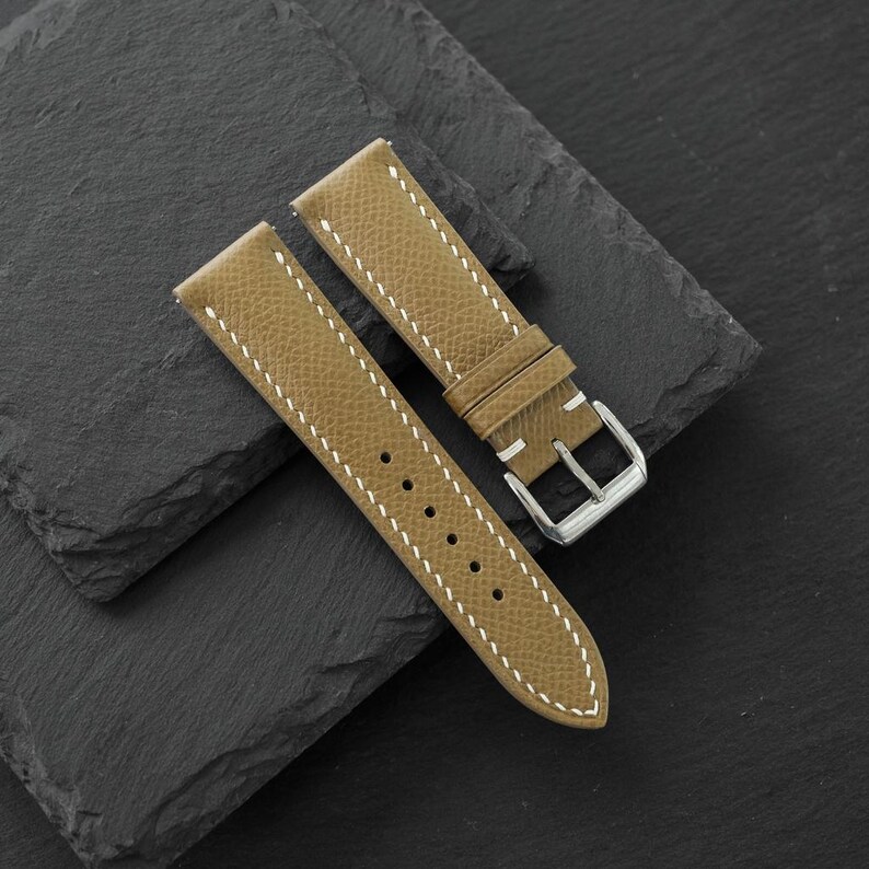 May include: A tan watch strap with a textured surface and white contrast stitching. The strap features a silver-toned metal buckle and two matching tan leather keepers. It has a series of circular holes for adjustment along the longer piece.