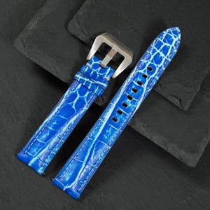 May include: A bright blue watch strap featuring a textured, embossed pattern resembling alligator skin. The strap includes a polished silver-tone metal buckle and a series of punched holes for adjustment, showcasing a glossy finish throughout.