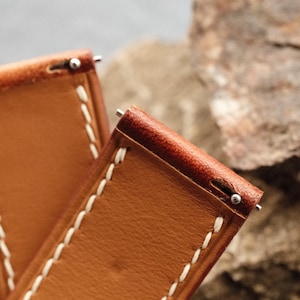 Pode incluir: Duas pulseiras de rel&oacute;gio em couro castanho-claro com costuras brancas vis&iacute;veis ao longo das bordas. Cada pulseira inclui um mecanismo de mola em metal prateado na extremidade superior para fixa&ccedil;&atilde;o &agrave; caixa do rel&oacute;gio, com textura lisa.