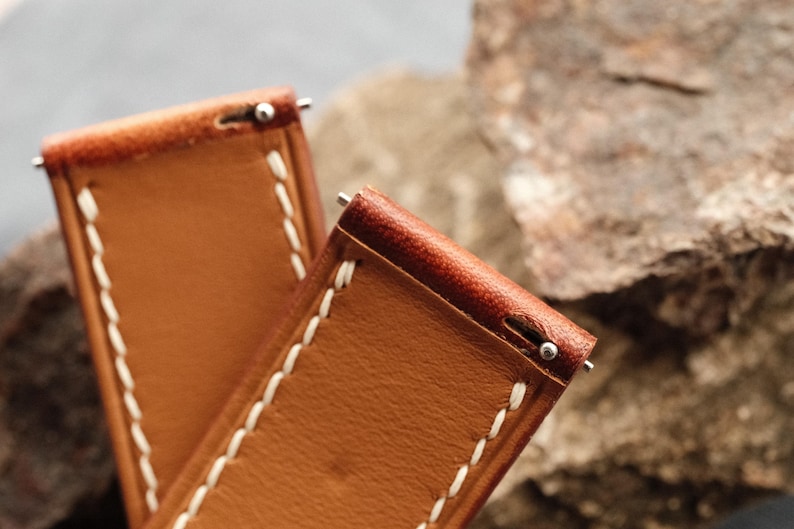 Pode incluir: Duas pulseiras de rel&oacute;gio em couro castanho-claro com costuras brancas vis&iacute;veis ao longo das bordas. Cada pulseira inclui um mecanismo de mola em metal prateado na extremidade superior para fixa&ccedil;&atilde;o &agrave; caixa do rel&oacute;gio, com textura lisa.