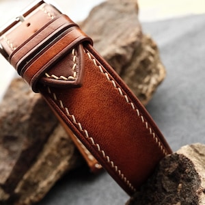 Pode incluir: Uma pulseira de rel&oacute;gio em couro castanho com um acabamento liso e polido, apresentando costuras vis&iacute;veis em cor creme ao longo das bordas. A pulseira inclui uma fivela de metal e dois passadores de couro, com um design c&oacute;nico e pontiagudo.