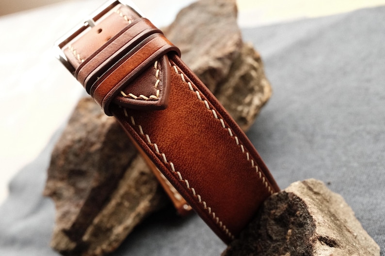 Pode incluir: Uma pulseira de rel&oacute;gio em couro castanho com um acabamento liso e polido, apresentando costuras vis&iacute;veis em cor creme ao longo das bordas. A pulseira inclui uma fivela de metal e dois passadores de couro, com um design c&oacute;nico e pontiagudo.