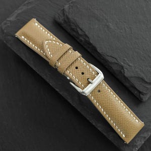 May include: A tan watch strap with a textured surface and white contrast stitching. The strap features a silver-toned metal buckle and a matching tan leather keeper loop, designed for securing a timepiece to a wrist with a classic pin closure.