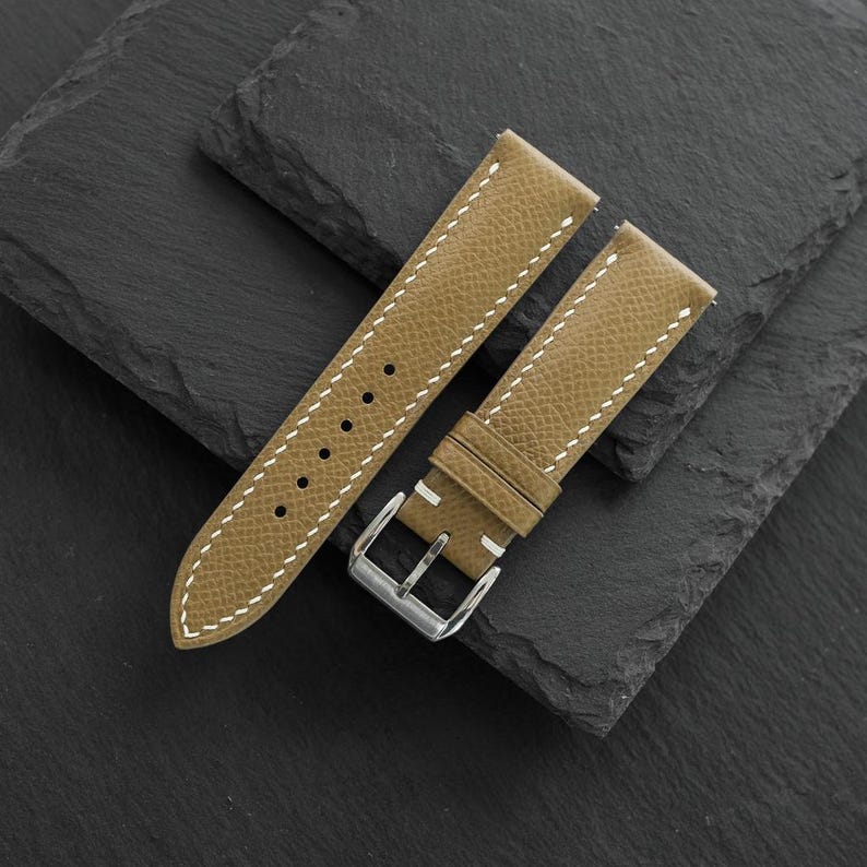 May include: A textured tan leather watch strap featuring white contrast stitching along the edges. The strap includes a silver-toned metal buckle and a series of circular adjustment holes for securing the watch to a wrist.