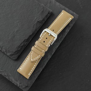 May include: A tan watch strap with a textured surface and white stitching. The strap features a silver-toned metal buckle and two matching tan leather keepers. The design includes multiple adjustment holes for a secure fit on a wrist.