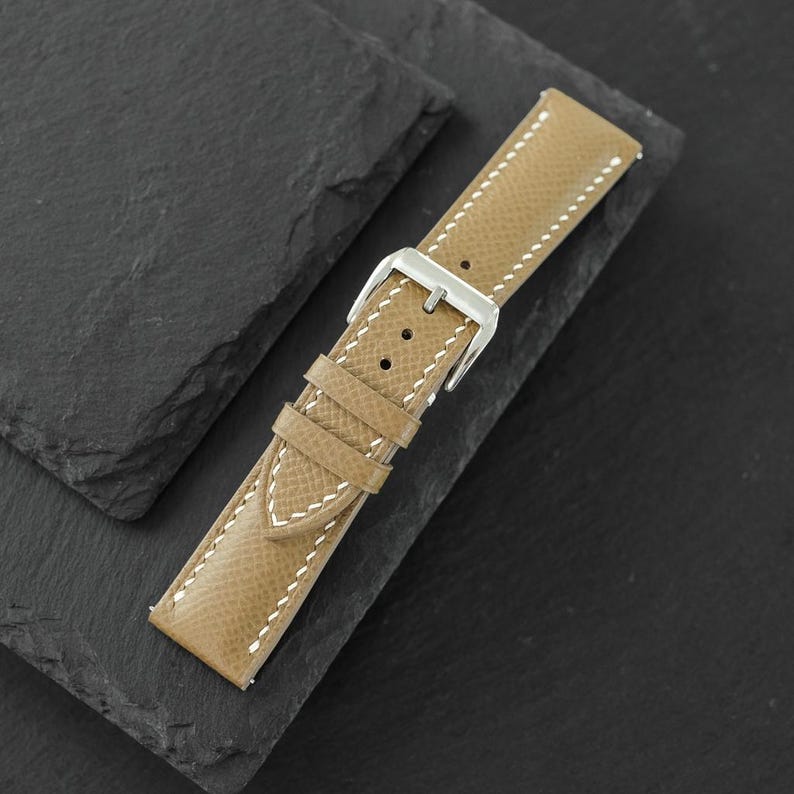 May include: A tan watch strap with a textured surface and white stitching. The strap features a silver-toned metal buckle and two matching tan leather keepers. The design includes multiple adjustment holes for a secure fit on a wrist.