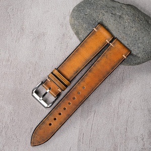 以下が含まれることがあります： シルバーのバックルが付いた、使い古した茶色の革製腕時計ストラップ。 ストラップにはステッチが見え、ヴィンテージまたは素朴なスタイルを示唆する、使い古された外観です。 ストラップは、灰色の表面の隣にある灰色の石の上に置かれています。