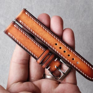 Pode incluir: Uma bracelete de rel&oacute;gio em pele castanho-clara de duas pe&ccedil;as com um acabamento degrad&eacute; que transita de um centro mais claro para bordas mais escuras. A bracelete inclui costuras brancas em contraste, uma fivela de metal prateado e cinco furos.