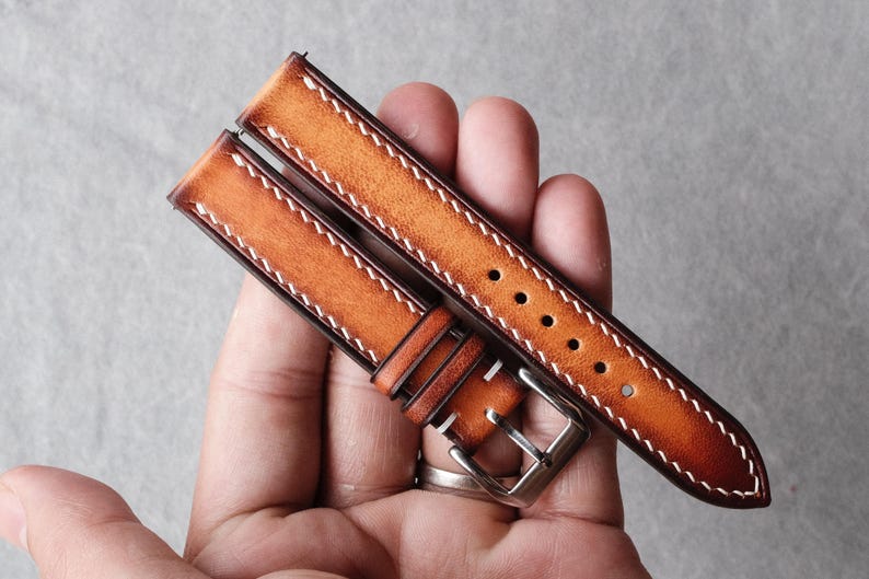 Pode incluir: Uma bracelete de rel&oacute;gio em pele castanho-clara de duas pe&ccedil;as com um acabamento degrad&eacute; que transita de um centro mais claro para bordas mais escuras. A bracelete inclui costuras brancas em contraste, uma fivela de metal prateado e cinco furos.