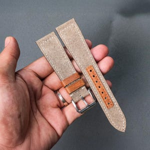 May include: Two-piece watch strap set. The straps are a light tan canvas material with white stitching. Brown leather accents are visible near the buckle and on the strap with the adjustment holes. A silver-coloured buckle is also present.
