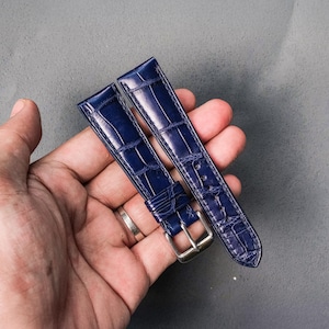 Op de afbeelding: Twee marineblauwe horlogebandjes met een krokodillenpatroon. De bandjes hebben zilverkleurige gespen en worden in een hand gehouden tegen een grijze achtergrond. De bandjes zijn ongeveer 23 cm lang.