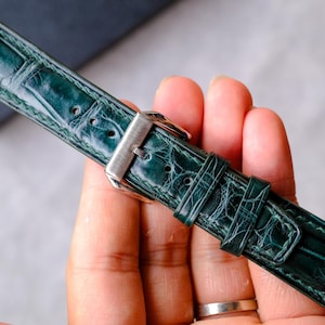 Op de afbeelding: Een donkergroene horlogeband met een glanzende, getextureerde afwerking die een reptielhuidpatroon nabootst. De band is voorzien van een gepolijste zilverkleurige metalen gesp en twee bijpassende lederen lussen voor een veilige bevestiging.