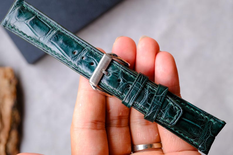 Op de afbeelding: Een donkergroene horlogeband met een glanzende, getextureerde afwerking die een reptielhuidpatroon nabootst. De band is voorzien van een gepolijste zilverkleurige metalen gesp en twee bijpassende lederen lussen voor een veilige bevestiging.