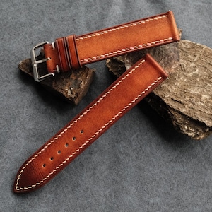 Pode incluir: Uma bracelete de rel&oacute;gio de duas pe&ccedil;as feita de couro castanho com acabamento em gradiente, apresentando costuras brancas em contraste ao longo das bordas. A bracelete inclui uma fivela de metal prateado e dois passadores de couro.