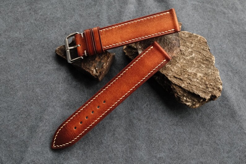 Pode incluir: Uma bracelete de rel&oacute;gio de duas pe&ccedil;as feita de couro castanho com acabamento em gradiente, apresentando costuras brancas em contraste ao longo das bordas. A bracelete inclui uma fivela de metal prateado e dois passadores de couro.