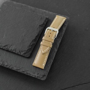May include: A tan watch strap with a textured surface and white contrast stitching. The strap features a silver-toned metal buckle and two matching tan leather keepers, designed for securing a timepiece to a wrist with a classic pin closure.