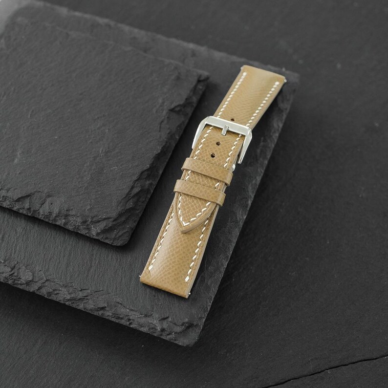 May include: A tan watch strap with a textured surface and white contrast stitching. The strap features a silver-toned metal buckle and two matching tan leather keepers, designed for securing a timepiece to a wrist with a classic pin closure.