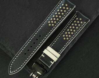 May include: A black leather watch strap featuring a perforated design with a light beige underside visible through the holes. The strap includes white contrast stitching along the edges, two matching leather keepers, and a black metal deployment clasp.