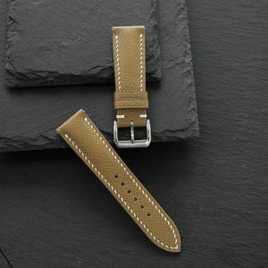May include: A tan watch strap with a textured surface and white contrast stitching. The strap features a silver-toned metal buckle and a matching tan leather keeper loop. The two-piece band includes a series of punched holes for adjustment.