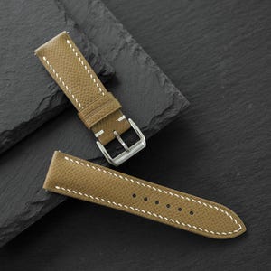 May include: A tan watch strap with a textured surface and white contrast stitching. The strap features a silver-toned metal buckle and a series of circular holes for adjustment, showcasing a classic design suitable for various timepieces.
