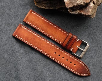 Può includere: Un cinturino per orologio in due pezzi realizzato in pelle color cuoio con una finitura liscia e opaca. Il cinturino presenta cuciture a contrasto chiare lungo i bordi, una serie di fori di regolazione circolari e una fibbia in metallo argentato.