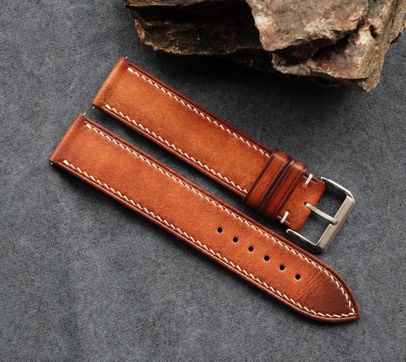 Pode incluir: Uma bracelete de rel&oacute;gio de duas pe&ccedil;as feita de couro castanho-claro com um acabamento liso e mate. A bracelete apresenta costuras de contraste claras ao longo das bordas, uma s&eacute;rie de orif&iacute;cios de ajuste circulares e uma fivela de metal prateado.